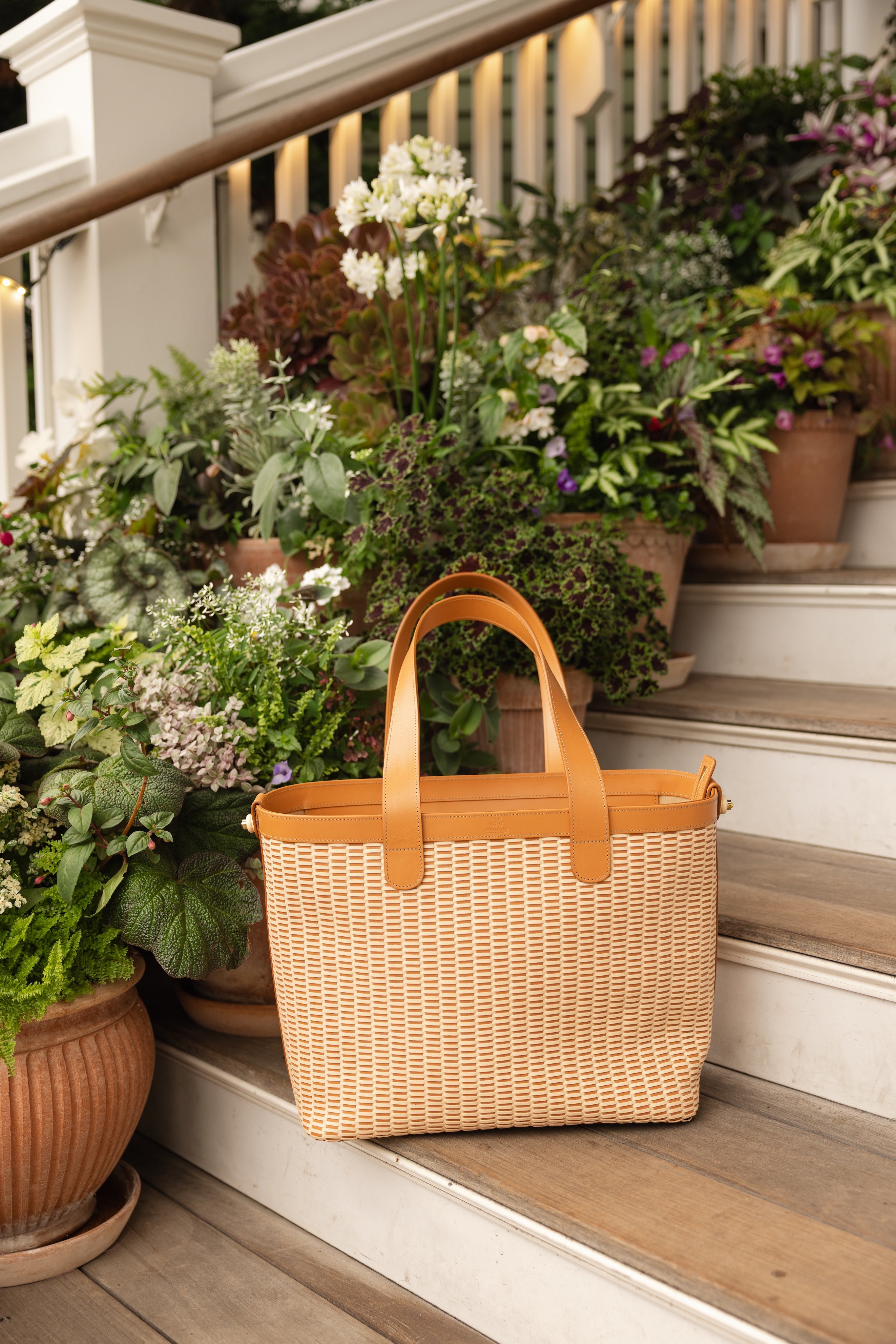 A woven straw tote bag with tan leather handles sits on wooden porch steps surrounded by potted green plants.