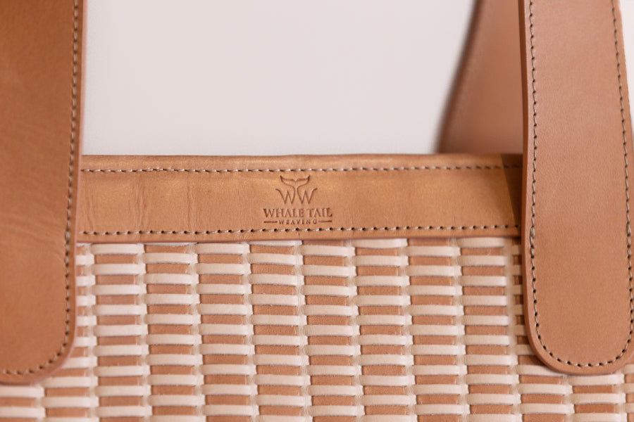 Close-up of a woven tote bag with tan leather trim and handles, showing the embossed Whale Tail Weaving Handbags logo on the leather band.