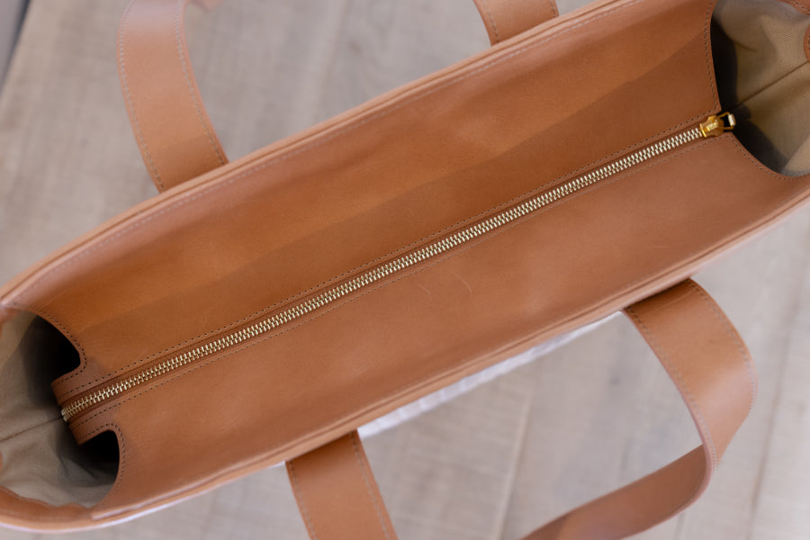 Close-up of the interior of a tan leather tote bag, showing a gold-tone zipper running down the center and matching leather handles on a light wood floor.