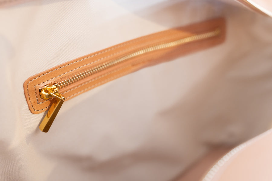 Close-up of a light beige or cream fabric bag interior with a tan leather zipper trim and gold-tone metal zipper pull.