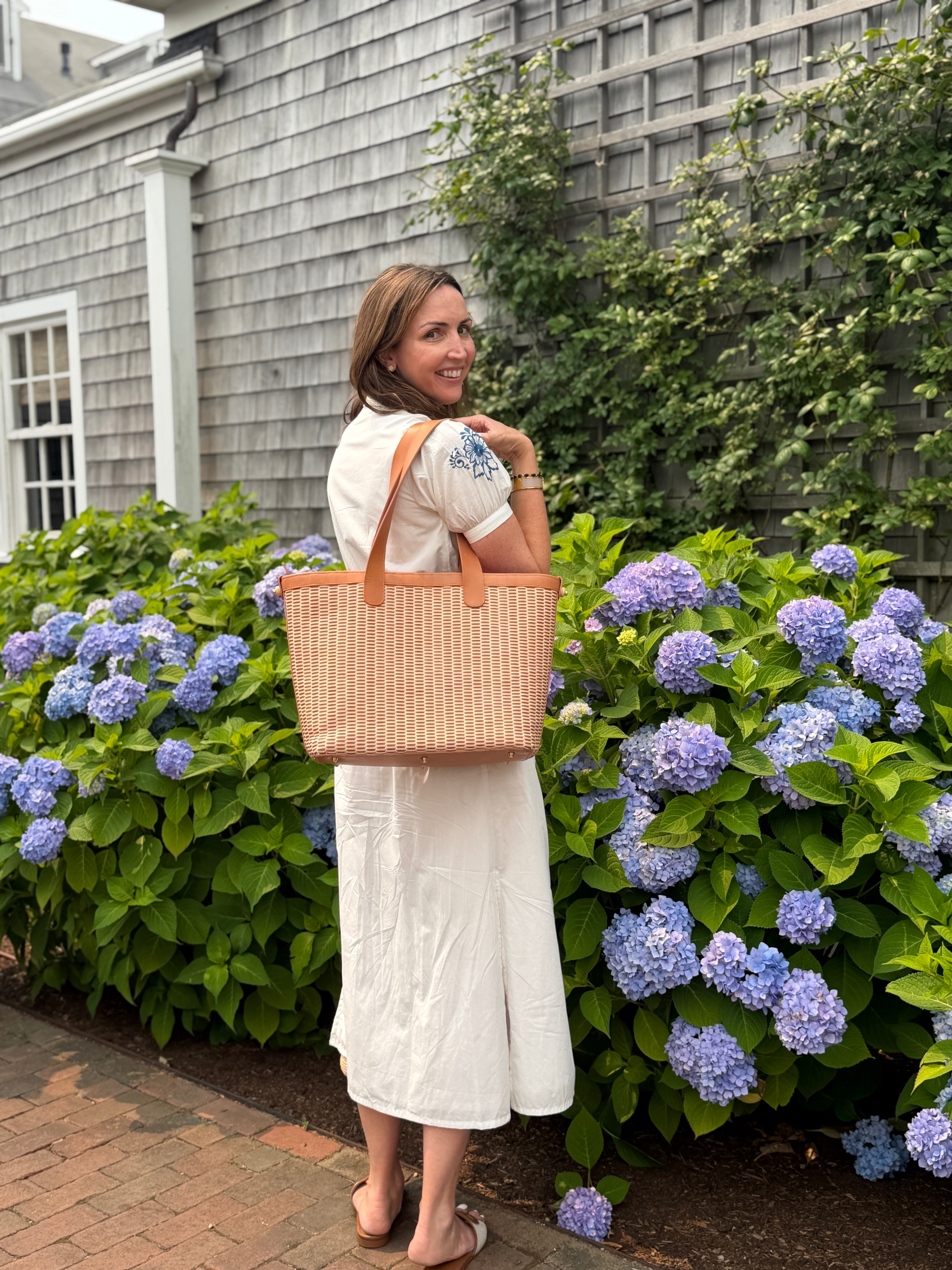 A woman in a white dress stands outdoors in front of hydrangea bushes, carrying a large woven Whale Tail Weaving tote bag with tan handles.