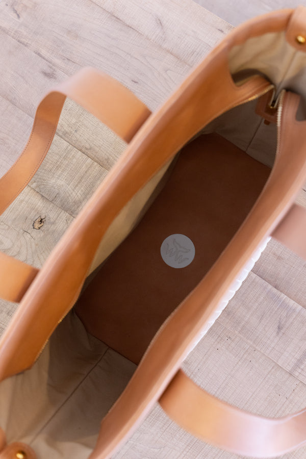 An overhead close-up of a large tote bag made of light tan leather, showing its interior lining and a circular logo medallion on the brown inner base, set against a light wood plank background.